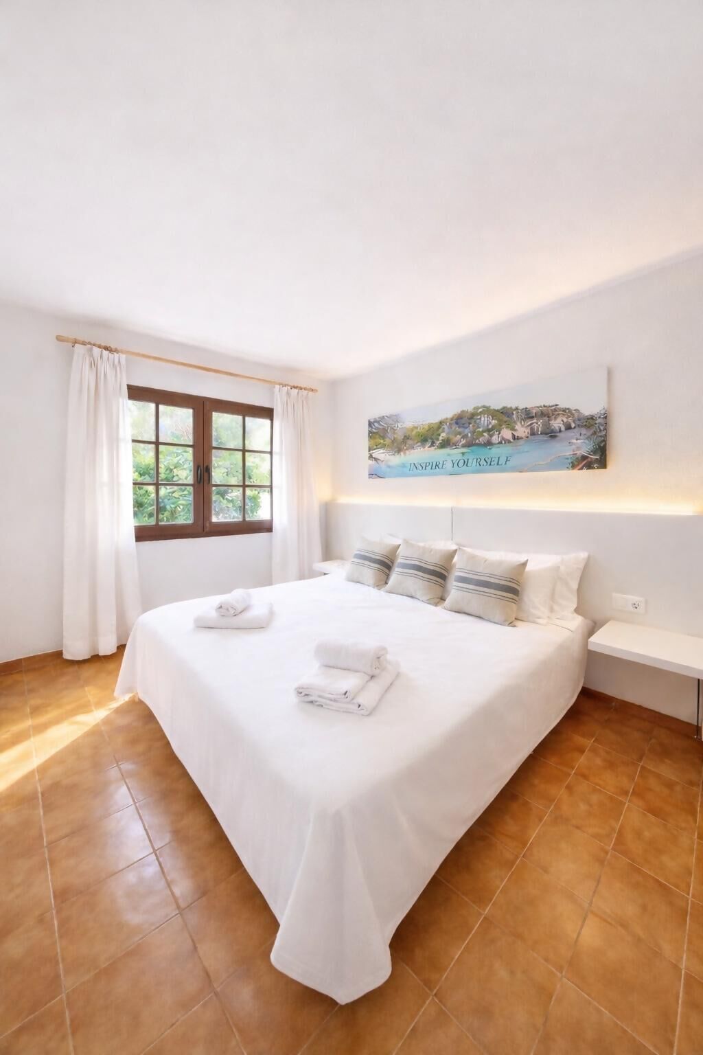 A neatly made bed with pillows and towels, a window with curtains, and a painting on the wall.