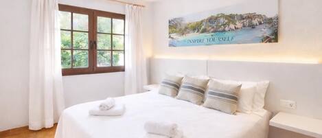 A neatly made bed with pillows and towels, a window with curtains, and a painting on the wall.