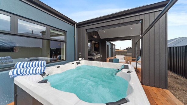 A modern outdoor hot tub area with a wooden deck, a covered seating area, and a view of the backyard. - CatchN'Relax with Hot Tub (Taupo)