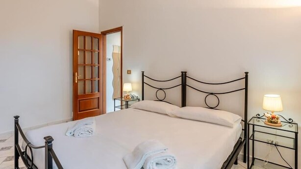Un lit bien fait, avec du linge de lit blanc et des serviettes pliées, une porte en bois avec des panneaux de verre, et une table de chevet avec une lampe.