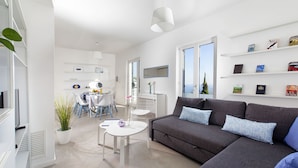 A modern living room with a grey sofa, white coffee table, and a dining area with chairs and a table. There are bookshelves with books and decorative items.