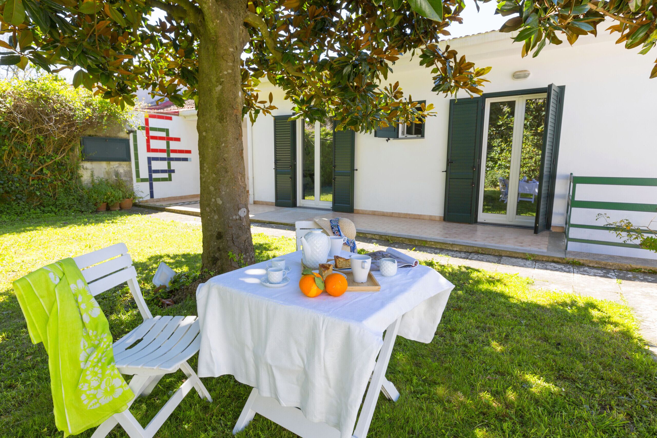 Una mesa al aire libre con mantel blanco, una silla y una bandeja con naranjas y tazas.