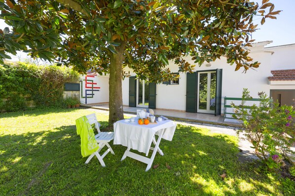 Une petite table d’extérieur, installée sur une pelouse verdoyante et recouverte d’une nappe blanche, est entourée d’un arbre aux feuilles vertes.