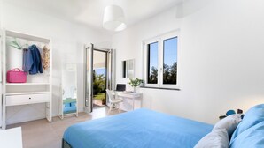 A bedroom with a bed, a desk, and a chair. There is a wardrobe with hanging clothes and a pink bag.