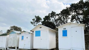 A row of white beach huts with blue shutters, numbered from 9 to 11, on a sandy beach with trees in the background. - Dimitri’s workshop 500m from the beach (Barbâtre)