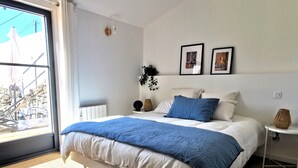 A bedroom with a large bed, a window with a view of a building, a small table with a lamp, and a wall-mounted light fixture. - Dimitri’s workshop 500m from the beach (Barbâtre)