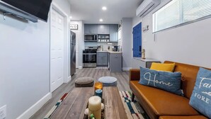 Een moderne woonkamer met een bank, een poef en een salontafel. In de achtergrond bevindt zich een keuken met apparatuur en kasten.