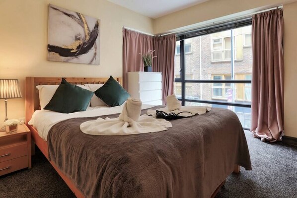 A hotel room with a large bed, a bedside table with a lamp, a dresser, and a window with curtains. - Lovely One Bed Apartment in Hip End of Temple Bar (Dublin 8)