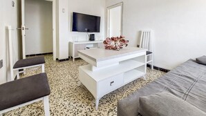 Een hotelkamer met een wit bureau, een televisie, een decoratieve bloem, een grijze bank en een vloer met een patroon.