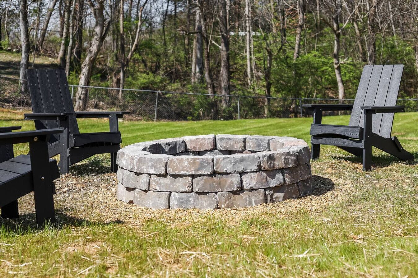 A circular stone fire pit surrounded by two black Adirondack chairs in a grassy outdoor area with trees in the background.