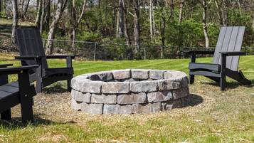 A circular stone fire pit surrounded by two black Adirondack chairs in a grassy outdoor area with trees in the background.