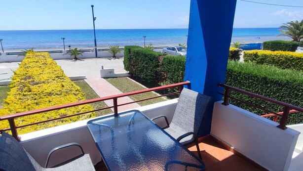 A balcony with a glass table and chairs, overlooking a landscaped area with yellow flowers and a view of the sea.