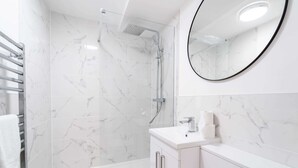 A modern bathroom with a glass-enclosed shower, a white marble-tiled wall, a round mirror, and a white sink with a countertop. - Central Chester: Cozy and Modern Flat (Chester)