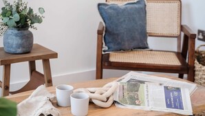 Une table basse en bois sur laquelle se trouvent un journal, deux tasses blanches, une plante et une chaise en osier avec un coussin bleu.