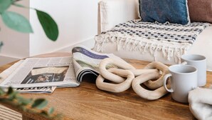 Un coin lecture confortable avec un fauteuil, une couverture, un journal, une paire d’anneaux en bois et deux tasses blanches.