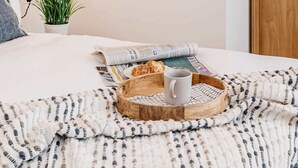 A neatly arranged bedroom with a bed, a bedside table with a lamp, framed abstract art, a small potted plant, and a tray with a mug and breakfast items. - Stylish Chester City Apartment (Chester)