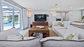 A modern living room with a large TV screen displaying 'iTrip Palm Harbor', a wooden coffee table with magazines, a grey sofa, and a view of a swimming pool through large windows. - Luxury 3BR Home w/ Heated Pool, Outdoor TV, Bikes & BBQ – Perfect for Families with Modern Kitchen! (Port Richey)