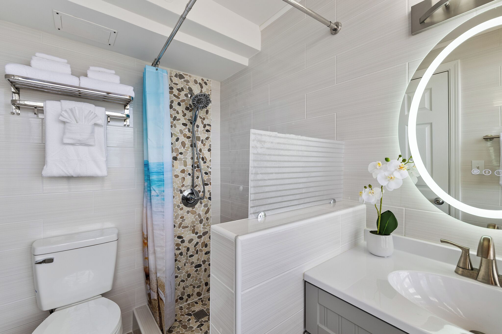 A modern bathroom with a white toilet, a shower with a mosaic tile wall, a white sink with a mirror, and a towel rack with folded towels.