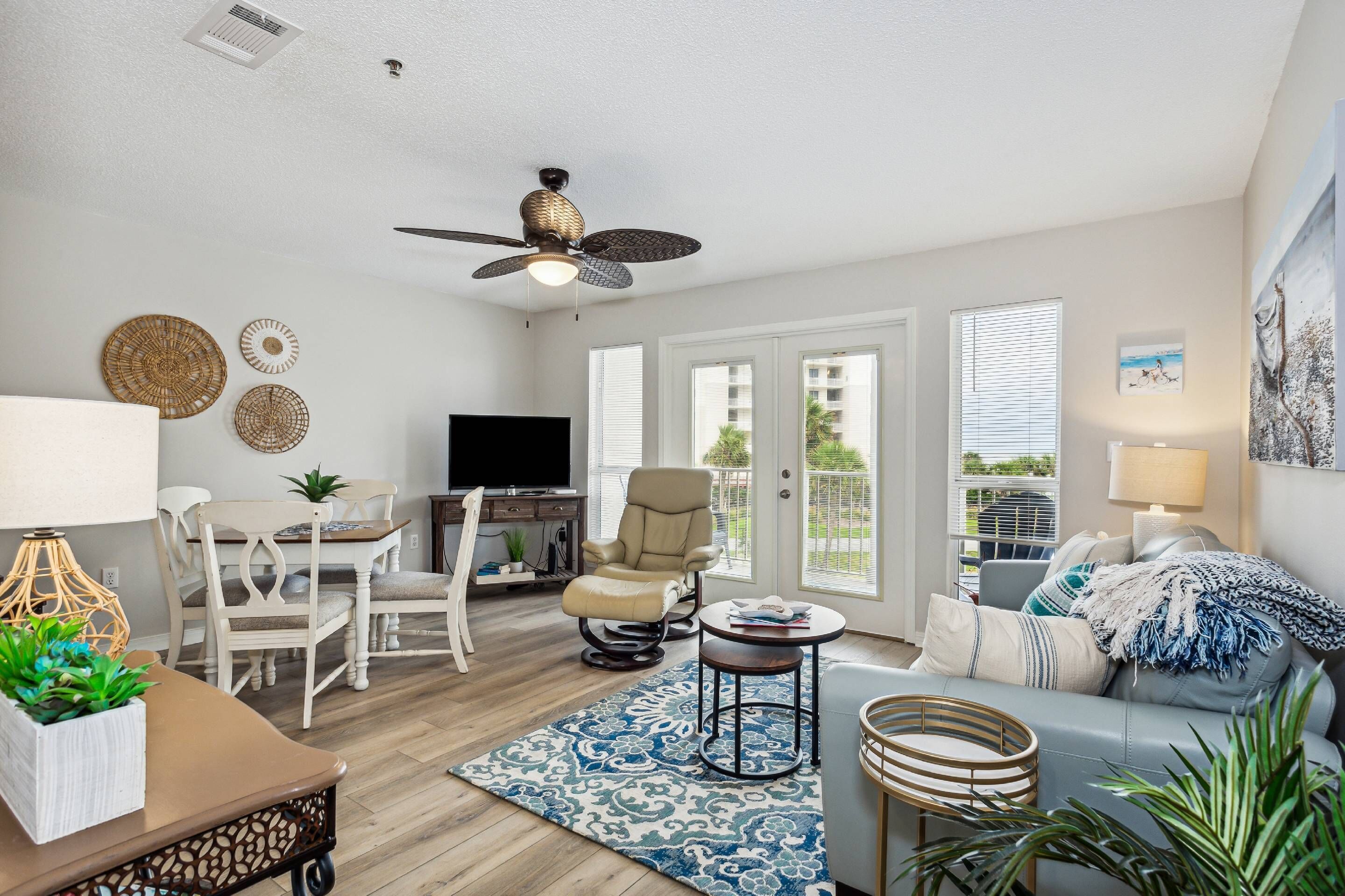 A spacious living room with a ceiling fan, a television, a dining area with chairs and a table, and a large window with blinds.