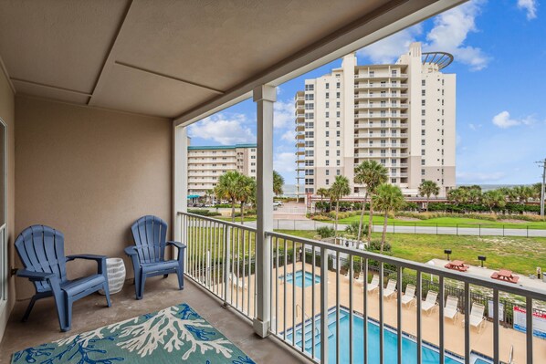 A balcony with two blue Adirondack chairs, a patterned rug, and a view of a swimming pool and tall buildings.