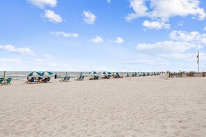 A beach with rows of blue and white beach chairs and umbrellas, a wooden fence, and a flag on a pole. - Completely Renovated 1BR Condo at Grand Caribbean West |Steps Away from the Gulf |Snowbird Friendly (Pensacola)