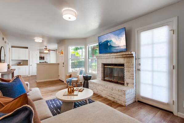 A spacious living room with a fireplace, a television mounted above it, and a kitchen in the background.