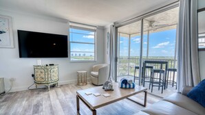 A living room with a flat-screen TV, a sofa, a coffee table, and a view of a balcony with outdoor seating.