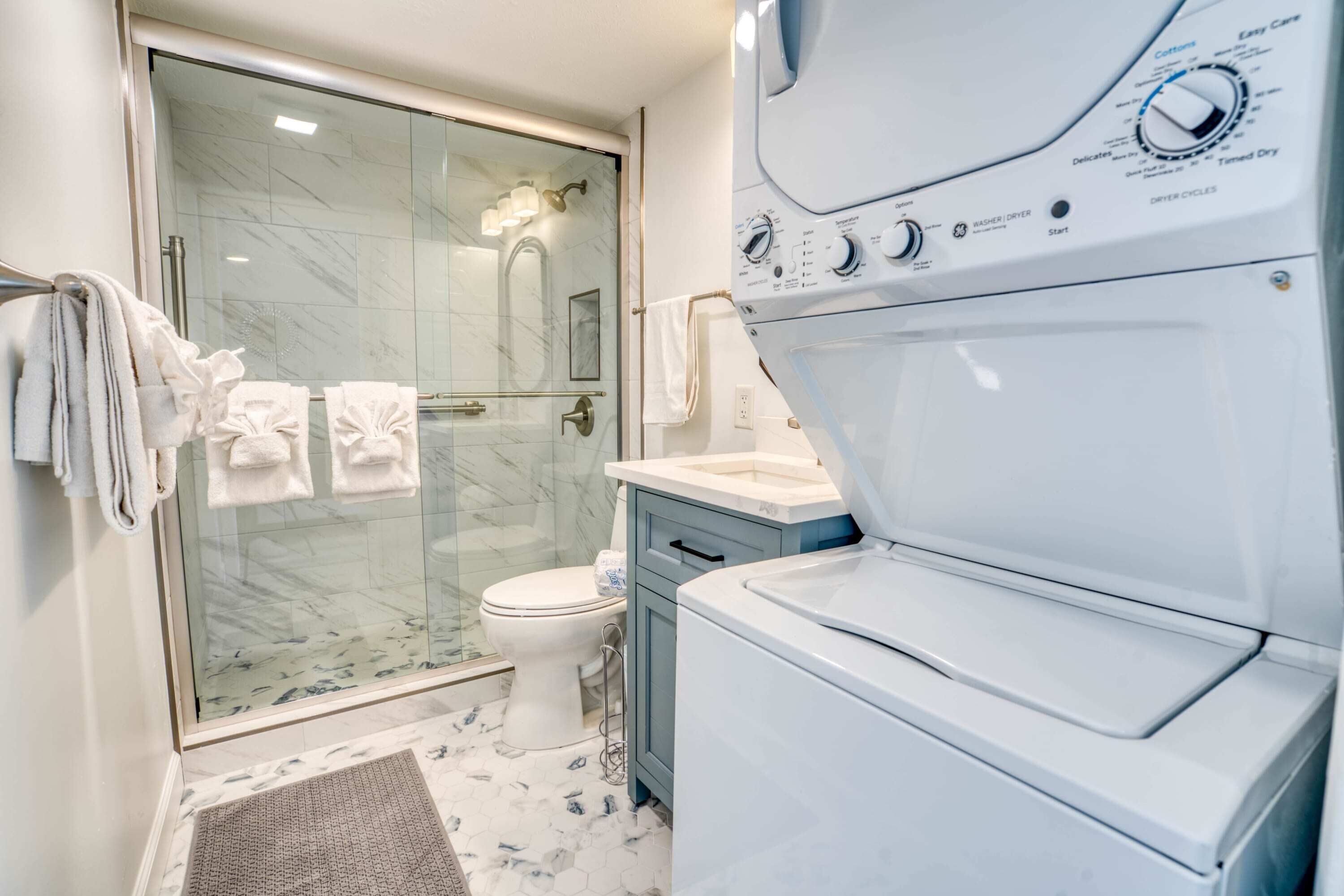 A modern bathroom with a glass shower enclosure, a white toilet, and a stacked washer and dryer unit.