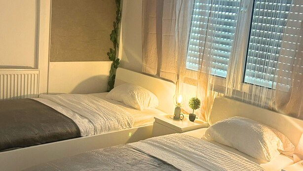 A double bed with a headboard, a bedside table with a lamp, and a window with blinds. - Stadtnahe Wohnung mit Küche - Ideal für Familien, Singles und Geschäftsreisende! (Oelde)