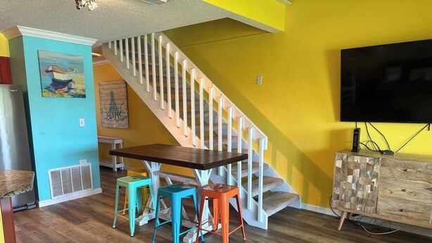 A living room with a wooden staircase, a dining table with colorful stools, a TV, and a wooden cabinet. - 10F, Wave Hello (Miramar Beach)