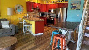 A modern kitchen with red cabinets, a central island, stainless steel appliances, and a dining area with a wooden table and bar stools. - 10F, Wave Hello (Miramar Beach)