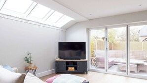 A modern living room with a flat-screen TV on a wooden stand, a sofa with cushions, a small side table with a plant, and a sliding glass door leading to an outdoor patio area. - Luxury Chichester Home for 12 (West Sussex)