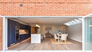 A modern kitchen with dark cabinetry, a central island, and a dining area with a table and chairs. - Luxury Chichester Home for 12 (West Sussex)