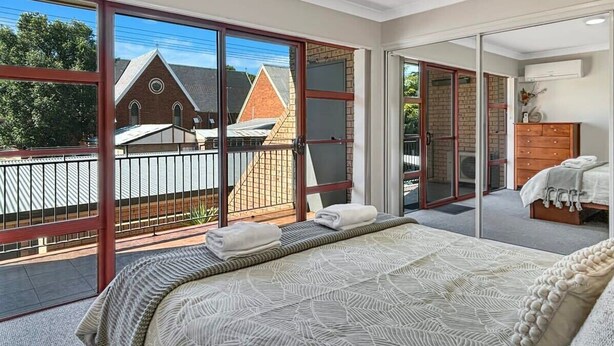 A bedroom with a large bed, a dresser, and a view of a brick building through large sliding glass doors. - Glenmore Lodge - CBD Location, Hidden Gem (Dubbo)