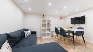 A modern living room with a dark grey sofa, a white shelving unit, a dining table with chairs, and a flat-screen TV mounted on the wall. - Sous Plex palais des congres Porte Maillot - 4P (Neuilly-sur-Seine)