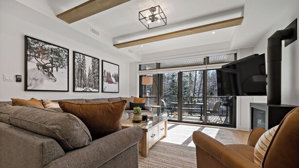 Una sala de estar moderna con un sofá gris grande, un sillón marrón y una mesa de centro. Hay dos cuadros enmarcados en la pared, una chimenea y un ventanal con vistas a árboles.