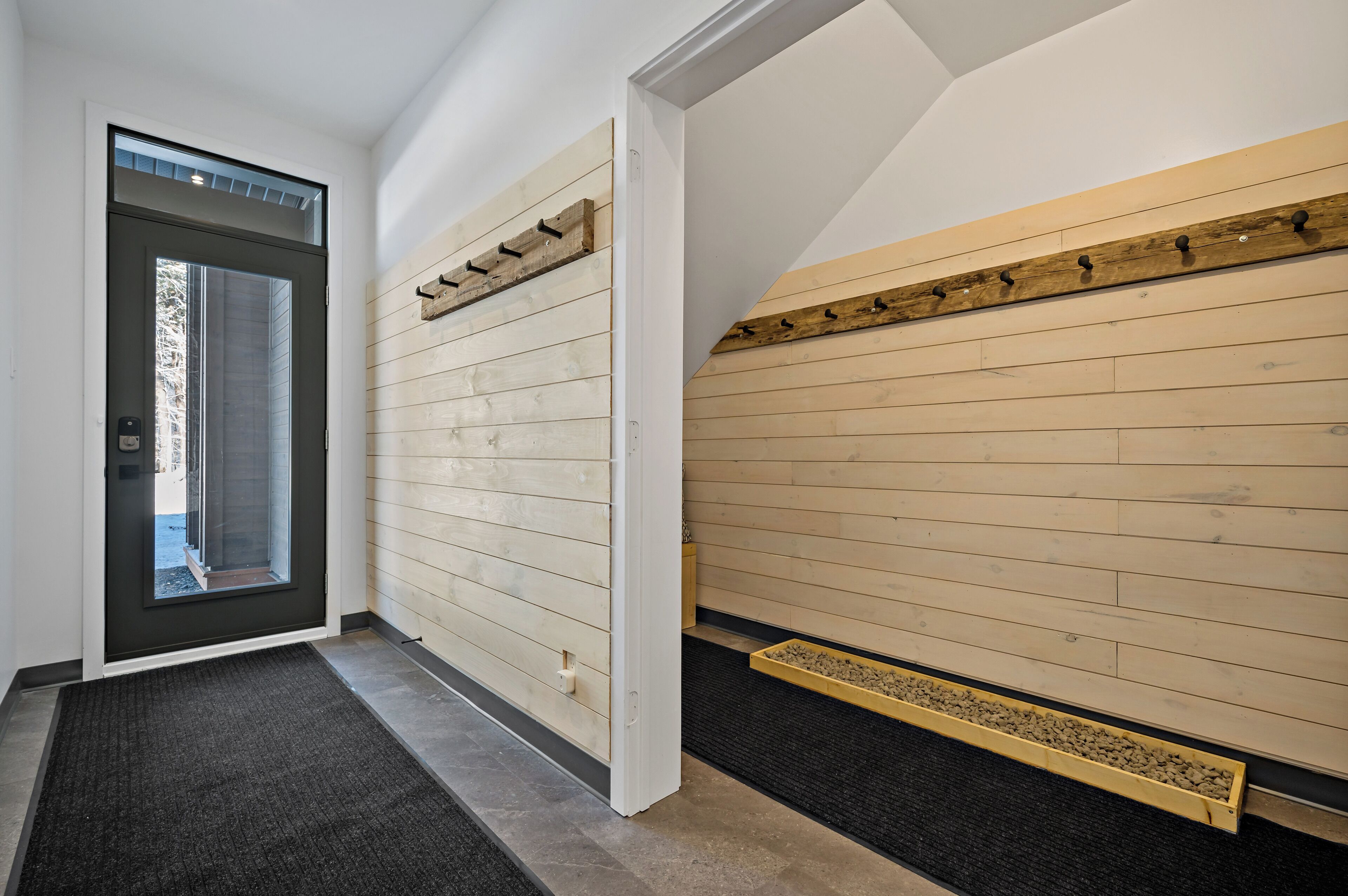 Un couloir avec une porte sombre, des murs recouverts de lambris en bois, un tapis noir et un petit bac à plantes rempli de gravier.