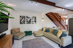A living room with a sectional sofa, a TV, a wooden cabinet, a staircase, and a wall with artwork. - The Surf Pad - Oceanstays - Pet Friendly (Angourie)