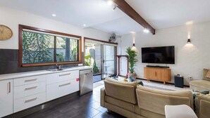 A modern kitchen with a large window, a dining area with a sofa, and a flat-screen TV mounted on a white brick wall. - The Surf Pad - Oceanstays - Pet Friendly (Angourie)
