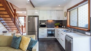 A modern kitchen with white cabinets, a stainless steel refrigerator, a dishwasher, and a large window with a brick wall view. - The Surf Pad - Oceanstays - Pet Friendly (Angourie)