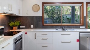 A modern kitchen with white cabinets, a built-in stove, a sink, and a window with a view of greenery. - The Surf Pad - Oceanstays - Pet Friendly (Angourie)