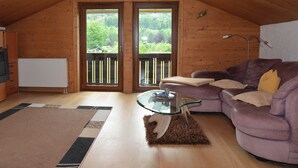 Een knus ingerichte kamer met houten wanden, een paarse bank, een glazen salontafel en uitzicht op groen.