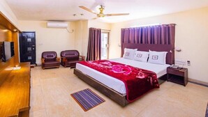 Quarto de hotel com uma cama, uma escrivaninha, duas cadeiras, um ventilador de teto e uma televisão.