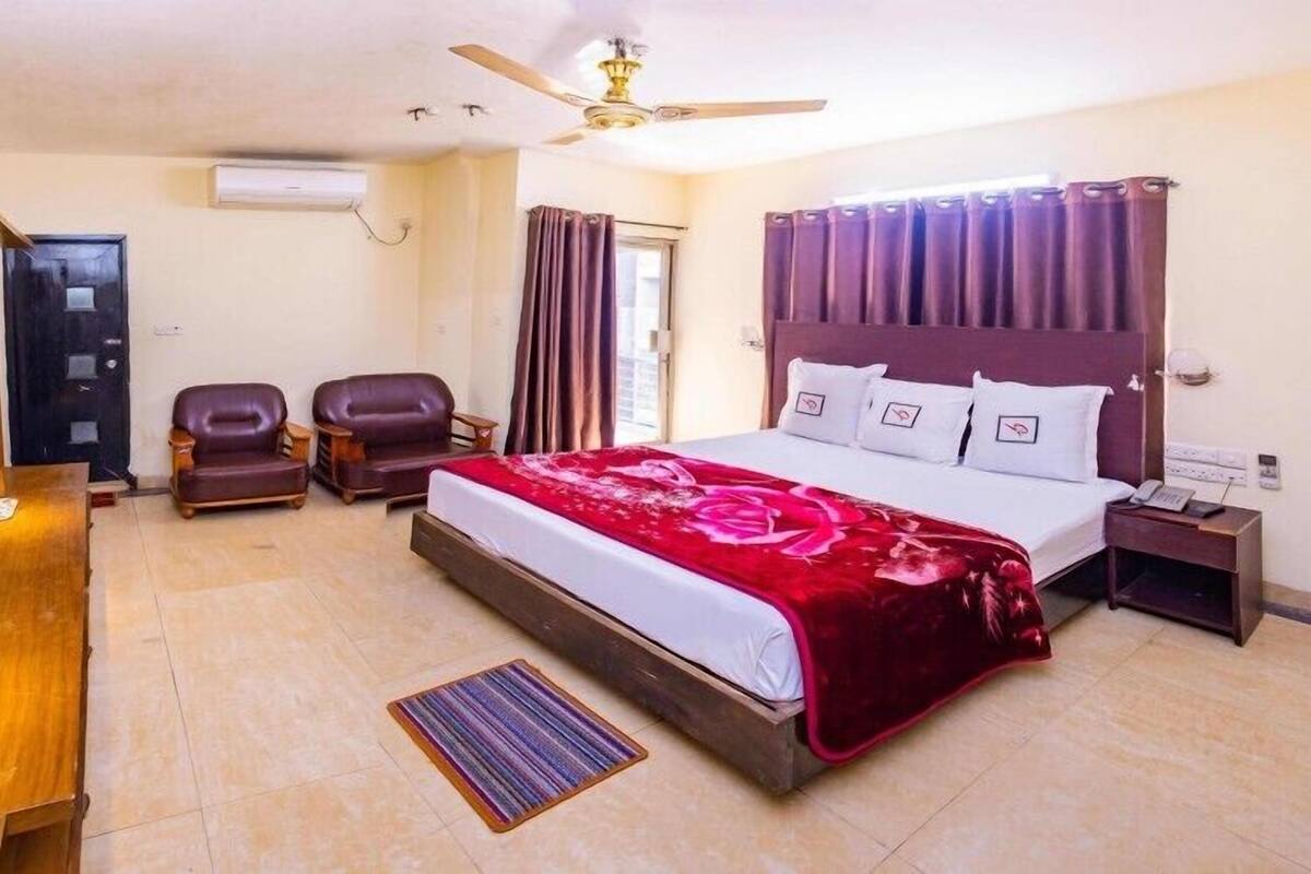 A hotel room with a bed, a desk, two chairs, a ceiling fan, and a TV.