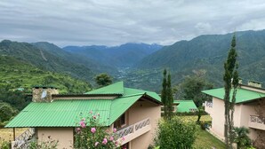 Une vue sur une vallée avec des bâtiments aux toits végétalisés, une structure en pierre surmontée d’une antenne parabolique, et une personne visible à travers une fenêtre.