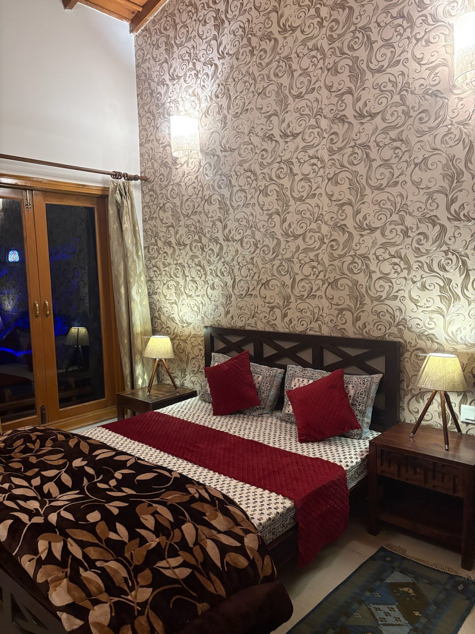 A bedroom with a bed, patterned wallpaper, a wooden headboard, two bedside tables with lamps, and a window with curtains.