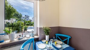 Una zona de menjador amb una taula preparada per a dues persones, una finestra amb vistes a vegetació i cases, i un vas amb flors al davant de la finestra.