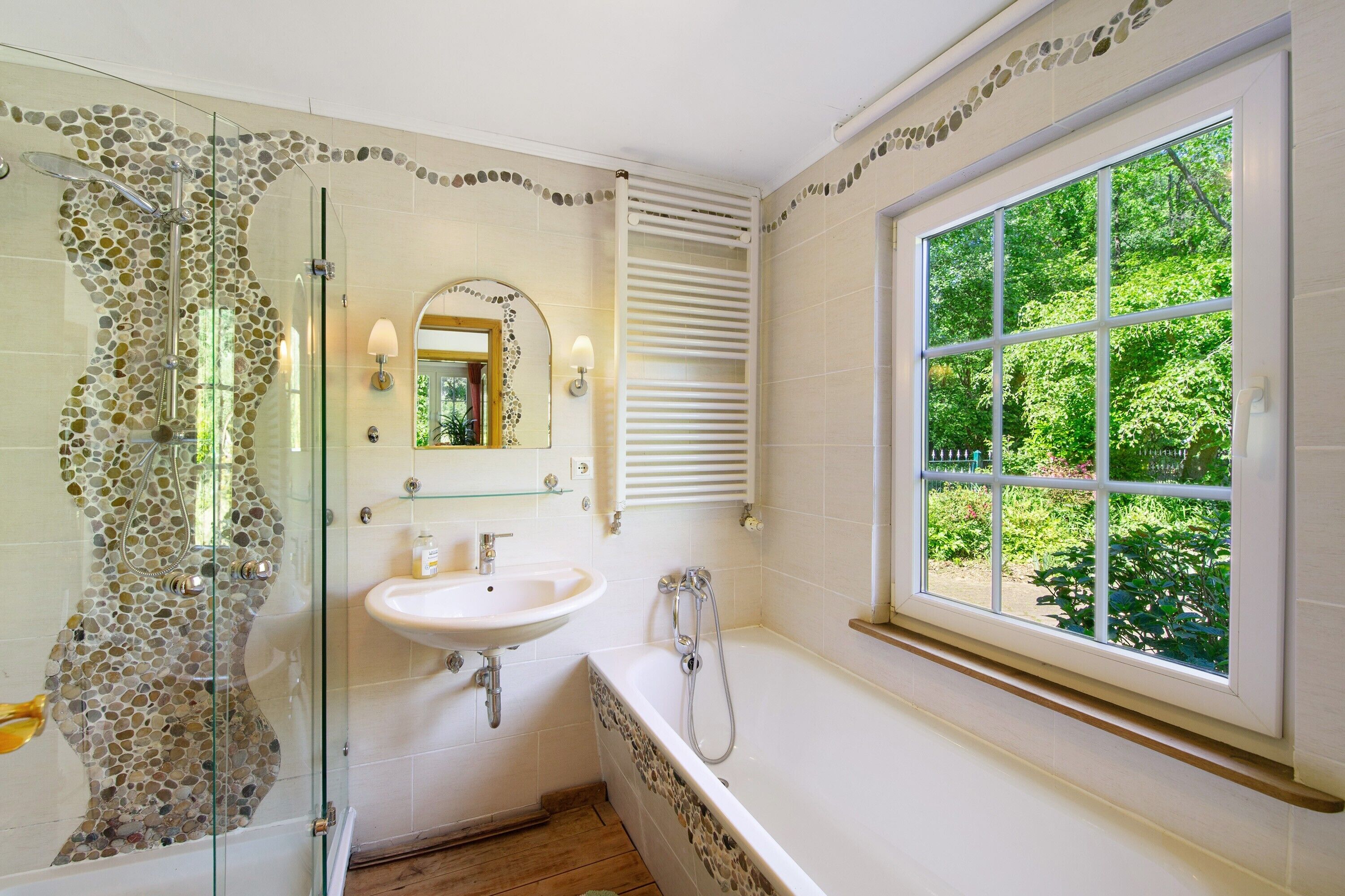 A bathroom with a glass shower, a sink, a bathtub, a window with a view of greenery, and a decorative wall.