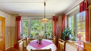 Une salle à manger avec une table, des chaises, un luminaire suspendu, des plantes en pot et de grandes fenêtres.