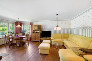 Ein Wohnzimmer mit einer Couch, einem Hocker, einem Esstisch, Stühlen, einem Fernseher und einem Schrank.
